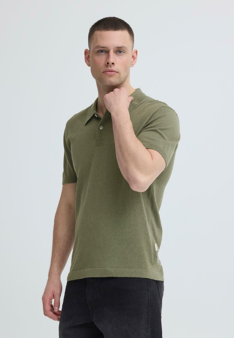 Jeune homme aux cheveux courts portant un polo vert olive et un pantalon noir, levant le poing près du menton, fond neutre.