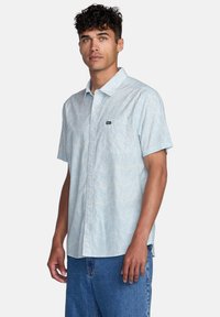 Short-sleeve, light blue button-up shirt with a subtle white pattern, featuring a chest pocket and a small logo patch. Cotton material.