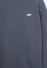 Felpa blu scuro realizzata in tessuto morbido, con un piccolo logo ricamato bianco sul lato sinistro del petto e dettagli di cucitura visibili sulle maniche.