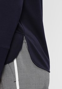 Námořnicky modrá mikina s oblým lemem a texturovaným materiálem. Kombinována se šedými kalhotami s károvaným vzorem, zdůrazňujícími kontrastní vzory.