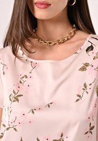 Blouse fleurie rose clair à manches courtes, avec des motifs complexes roses et verts. Collier chaîne en or en tant qu'accessoire principal.