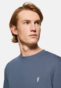 Lyseblå sweatshirt lavet af blødt stof. Har en rund halsudskæring og et lille hvidt logo på brystet. Glat tekstur og afslappet pasform.