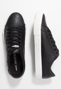 Mustat nahkakengät, joissa on sileä pinta, valkoiset kumipohjat, pyöreä kärki ja nauhakiinnitys. Sivussa ja kielessä on brändäystä.