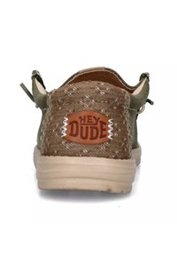 Zapato de tela verde con un tacón marrón texturizado, que presenta un parche con el logo "HEY DUDE". Suela de color beige claro, diseño sin cordones y con una lengüeta para facilitar el calzado.