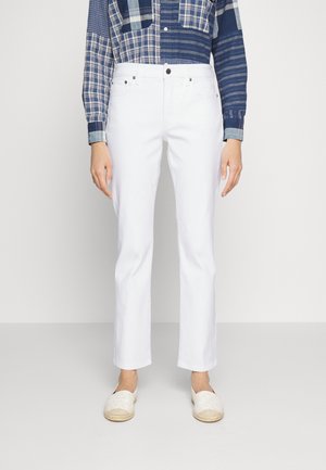 Person, die weiße gerade geschnittene Hose, ein blaues Patchwork-Karohemd und weiße Slipper trägt und vor einem schlichten Hintergrund steht.