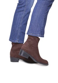 Botas de tobillo de ante marrón con un tac�ón bajo tipo bloque, que presentan una textura suave y un detalle de cremallera en el costado. Combinadas con unos vaqueros de denim azul.