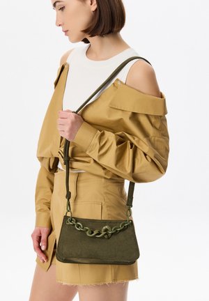 Borsa in suede verde oliva con tracolla a catena spessa, forma rettangolare e superficie liscia. La modella la tiene mentre indossa un giacchetto senape.
