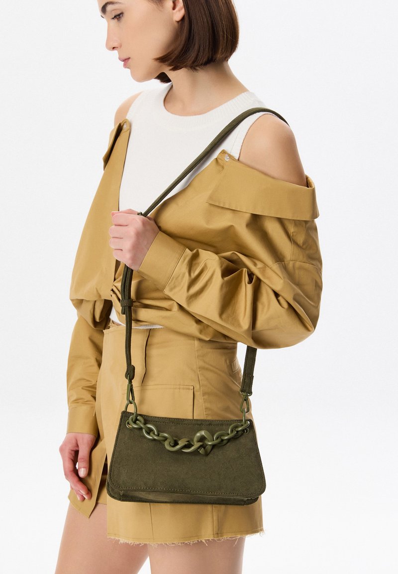 Borsa in suede verde oliva con tracolla a catena spessa, forma rettangolare e superficie liscia. La modella la tiene mentre indossa un giacchetto senape.
