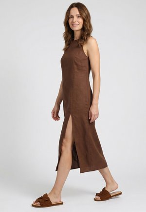 Vrouw met golvend bruin haar die een mouwloze bruine midi-jurk met een split aan de zijkant draagt, lopend op blote voeten in bruine instapsandalen met strikjes.