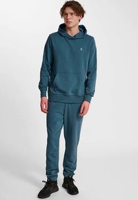 Set i teal med hoodie och joggers, med en känguruficka, ribbstickade muddar och en liten logga på bröstet. Mjukt tyg med ledig passform.