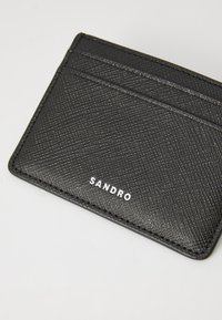 Tarjetero negro texturizado con cuatro ranuras para tarjetas, logo en relieve "SANDRO" en blanco y diseño rectangular y delgado. Material duradero.