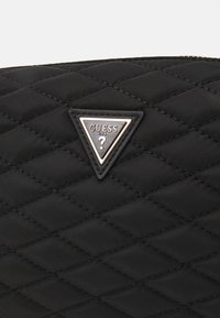 Gelaagd zwart stoffen tas met een driehoekig zilveren metalen logo-label met de tekst "GUESS". De textuur is glad met diamantpatronen.