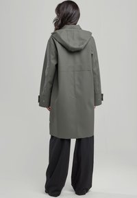 Vihreä vedenpitävä takki hupulla, jossa on suora leikkaus, pitkät hihat ja napilla suljettavat hihansuut. Silkkinen pinta ja minimalistinen muotoilu.