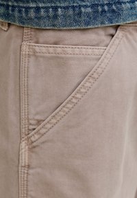 Gros plan sur la poche d'un pantalon beige avec des coutures visibles sous l'ourlet d'une veste en denim bleu.