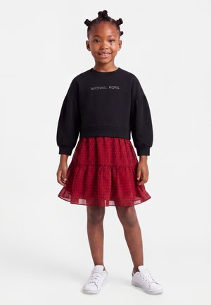 Ung pige med stylet hår iført en sort langærmet Michael Kors top, rød mønstret nederdel og hvide sneakers står mod en hvid baggrund.
