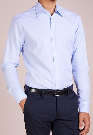 Mand iført lyseblå langærmet skjorte med knapper, stoppet ned i mørkeblå bukser med sort læderbælte, hænder afslappet ved siden.