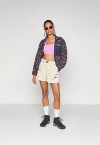 Πολύχρωμο μπουφάν με σχέδια, μοβ κοντό τοπ, μπεζ σορτς με τσέπες, λευκές κάλτσες και μαύρα μποτάκια. Τα γυαλιά ηλίου ολοκληρώνουν το look.