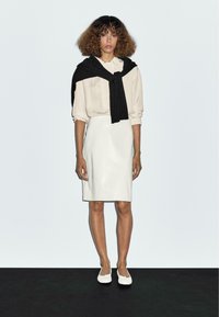 White leather pencil skirt paired with a light beige blouse. A black knitted sweater is worn casually over the shoulders. White flat shoes.