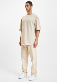 Beige oversized t-shirt med korte ærmer, parret med beige løse bukser og hvide sneakers, sat mod en simpel hvid baggrund.