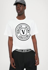 Baltā kokvilnas T-krekla ar melnu apļa Versace Jeans Couture logo, saskaņojumā ar melnām biksēm, kurām ir atbilstoša josta.