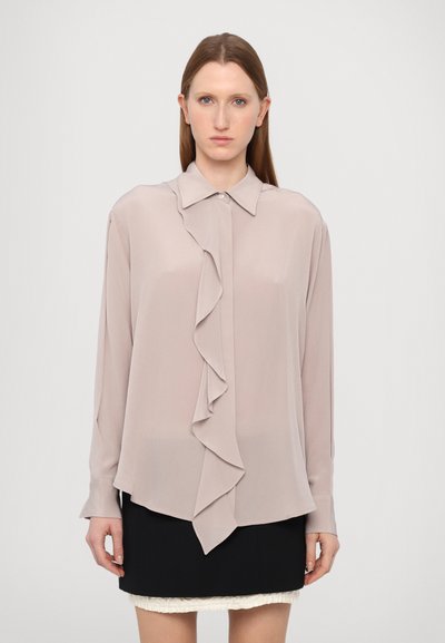 Blouse rose clair avec un devant à volants, manches longues, col et ourlet arrondi, associée à une jupe noire. Tissu lisse et léger.