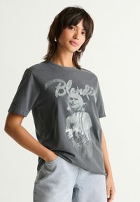Harmaa puuvillapaita, jossa on haalistunut graafinen printti henkilöstä ja teksti "Blondie." Lyhyet hihat ja pyöreä kaula-aukko.