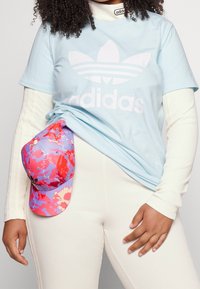 Ljusblå t-shirt med en vit Adidas-logotyp, tillsammans med en flerfärgad blommigare keps i rosa och lila. Modellen har på sig vita ribbade leggings.
