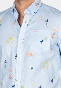 Lichtblauwe overhemden met een zak, bloemenpatronen in oranje, groen en blauw, en een linnenachtige textuur.