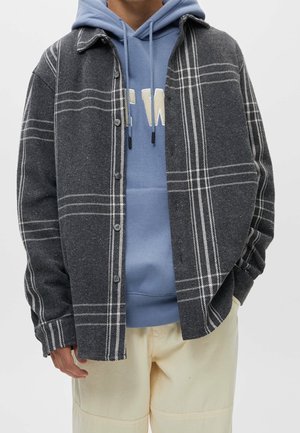 Persoon draagt een grijs-wit geruit overhemd met knopen over een blauwe hoodie met witte letters en lichtbeige broek, handen in de zakken.