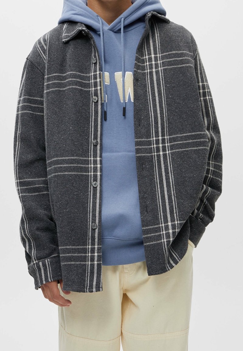 Personne portant une chemise à carreaux grise et blanche boutonnée sur un sweat à capuche bleu avec des lettres blanches et un pantalon beige clair, mains dans les poches.