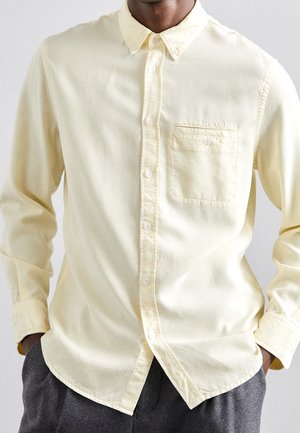 Homme portant une chemise boutonnée jaune pâle avec une poche poitrine et un pantalon gris foncé, mains dans les poches, sur un fond neutre.