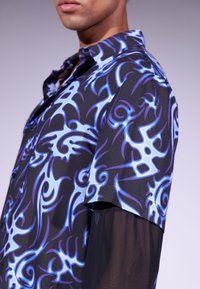 Camicia a maniche corte nera con intricati motivi tribali blu e bianchi. Presenta un colletto e un tessuto leggero e liscio.