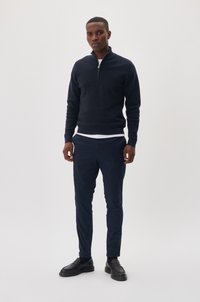 Pulover închiriat de marină din material moale, asortat cu pantaloni navy și mocasini negri. Maiou alb vizibil la guler.