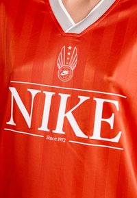 Tricou roșu Nike cu dungi verticale, având un logo alb și textul "Since 1972" pe față, și un guler în V cu margini gri.