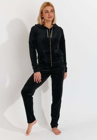 Tuta in velour nera con cappuccio zip e tasche frontali, abbinata a pantaloni. Texture morbida e design aderente.