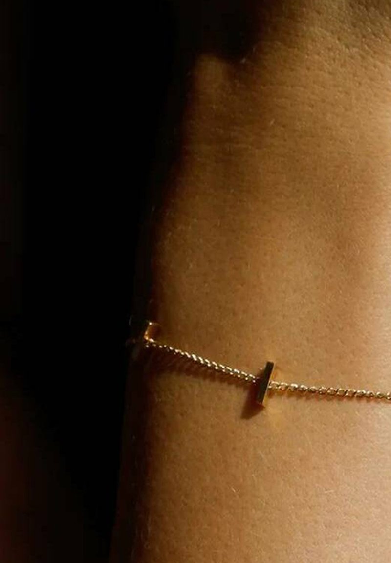 Goldene Kettenarmband mit einem minimalistischen, rechteckigen Anhänger und einem gedrehten Gliederdesign, das auf der Haut ruht und eine glatte Textur aufweist.