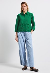 Chemise verte à boutons avec broderie en œillet à l'ourlet, associée à un pantalon en denim bleu clair à jambes larges et des chaussures en maille noires.