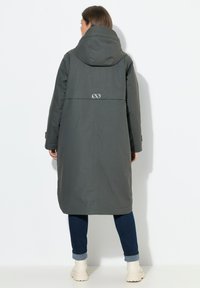 Chaqueta impermeable gris con capucha, diseño largo y un emblema en la parte posterior. Incorpora puños elásticos y un ajuste relajado. Llevada sobre unos vaqueros azules.