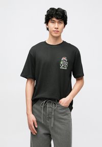 Tricou negru din bumbac cu un imprimeu colorat reprezentând un urs purtând o căciulă de Moș Crăciun, cu cuvântul „VISIONARY” dedesubt. Croială lejeră, mâneci scurte.