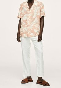 Homme portant une chemise à boutons à manches courtes à fleurs couleur pêche, un pantalon blanc et des sandales marron à enfiler sur un fond neutre.