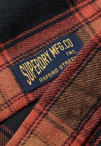 Punainen ja musta ruudullinen kangas, jossa on teksturoitu kudos. Sinisessä tarrassa lukee "SUPERDRY MFG. CO." kultaisella painatuksella.