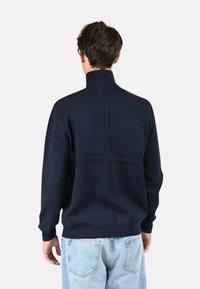 Pull bleu marine avec un col haut et un tissu texturé, présentant un dos sans couture et des poignets côtelés, assorti avec un jean bleu clair.