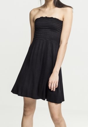 Vrouw draagt een zwarte mouwloze strapless jurk met een aansluitend, geplooid lijfje en een uitlopende rok, staand tegen een effen achtergrond.