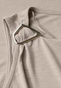 Top beige hecho de tejido texturizado con un detalle de diseño retorcido. Presenta un acento triangular metálico cerca del escote.