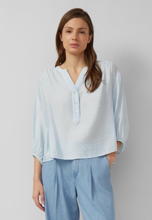 Femme aux longs cheveux bruns porte un chemisier bleu clair boutonné avec des manches 3/4 et un jean bleu clair ample, debout sur un fond uni.