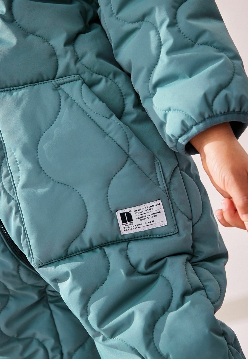 Combinaison matelassée bleu sarcelle avec une fermeture éclair à l'avant et une étiquette en patch sur la poche. La manche est dotée d'un poignet élastique pour un ajustement confortable.