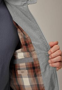 Chaqueta gris con una abertura con cremallera, que revela un forro interior a cuadros en tonos marrón y beige, y un bolsillo lateral con una etiqueta que dice "NEXT."