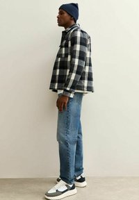 Man in een blauwe beanie, zwart-witte geblokte jas, blauwe jeans en witte sneakers die zijwaarts staat tegen een effen achtergrond.