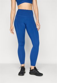 Leggings azuis de cintura alta com tecido liso e elástico. Apresentam bolsos laterais e costuras contrastantes, combinados com ténis desportivos pretos.