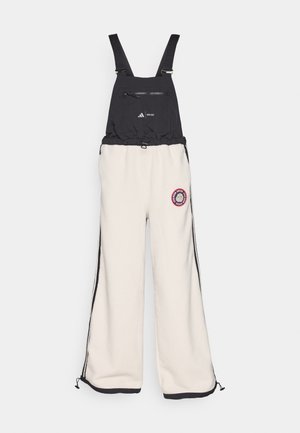 Schwarz-weißer Overall mit Fleece-Textur, ausgestattet mit einer Brusttasche mit Reißverschluss, verstellbaren Trägern und seitlichen Reißverschlüssen an den Hosenbeinen.